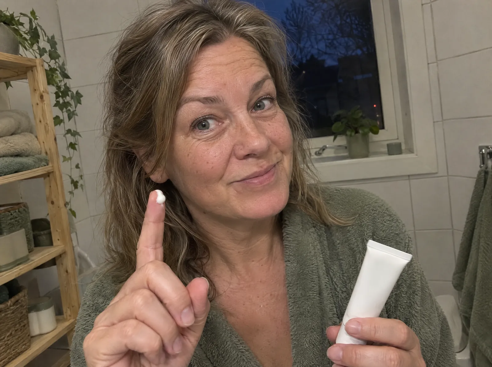 Kvinne i badekåpe holder opp pekefingeren med en ertestor dott krem, klar til å smøre den i ansiktet, og en merkeløs tube i den andre hånden.