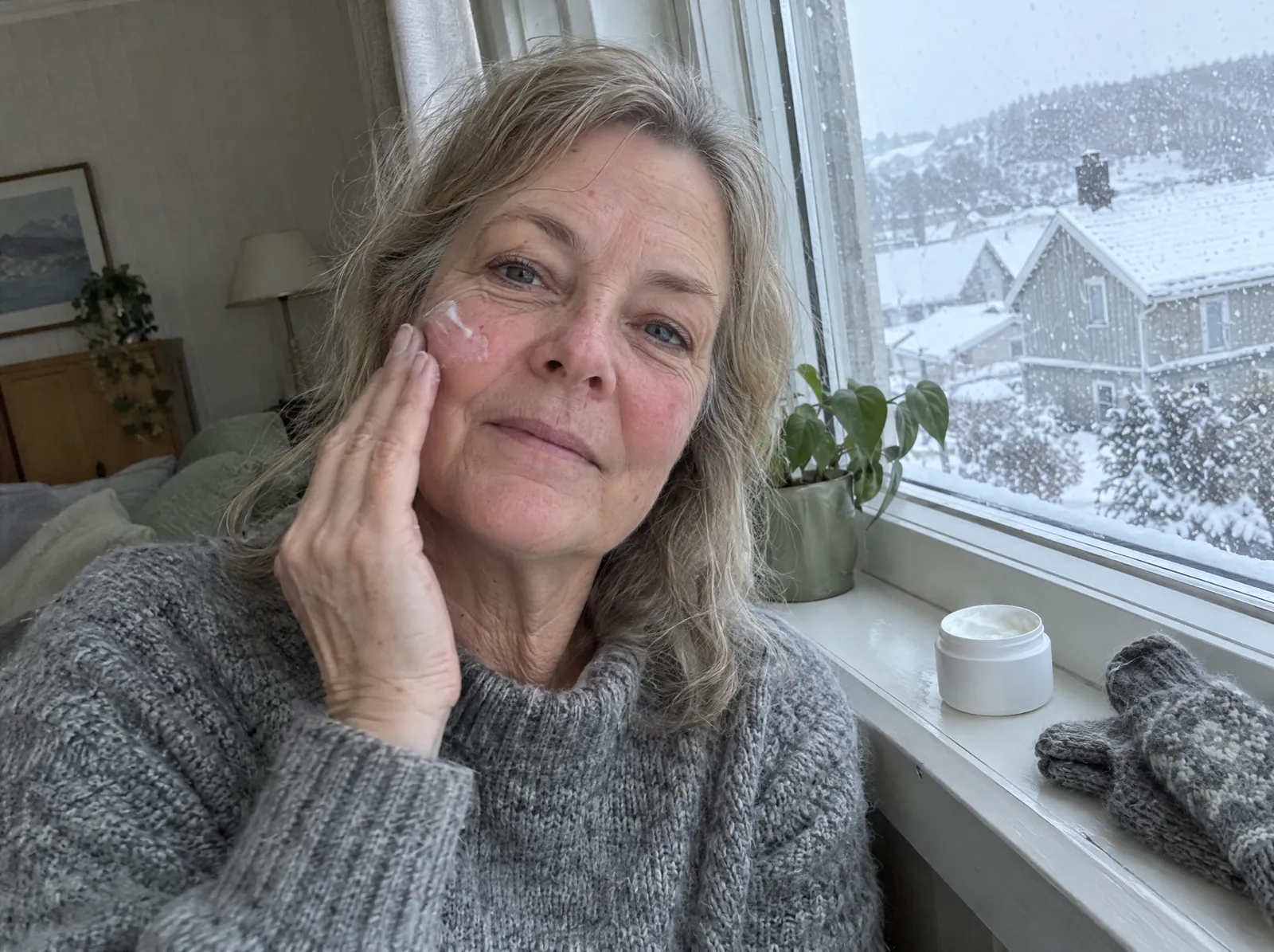 Kvinne i 60-årene i strikkegenser smører krem på kinnet ved et vindu der snøen laver ned, med en kremkrukke og ullvotter i vinduskarmen.